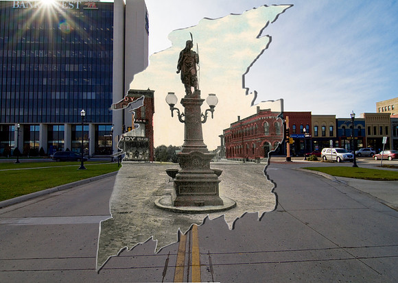 A statue of a Native American stands in a square in the business district. A horse and wagon is parked next to a utility pole at right. The first building on the right is the Commercial Bank, with Eme